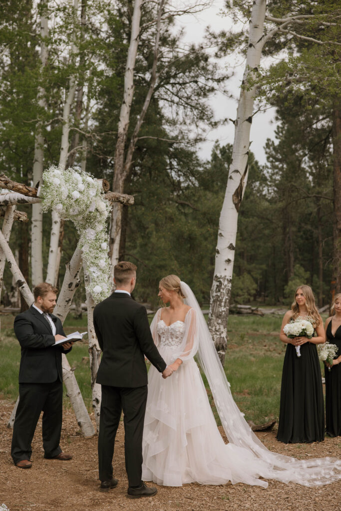 ceremony photo of groom holding hands at arbor with bride during outdoor wedding day at arizona nordic village