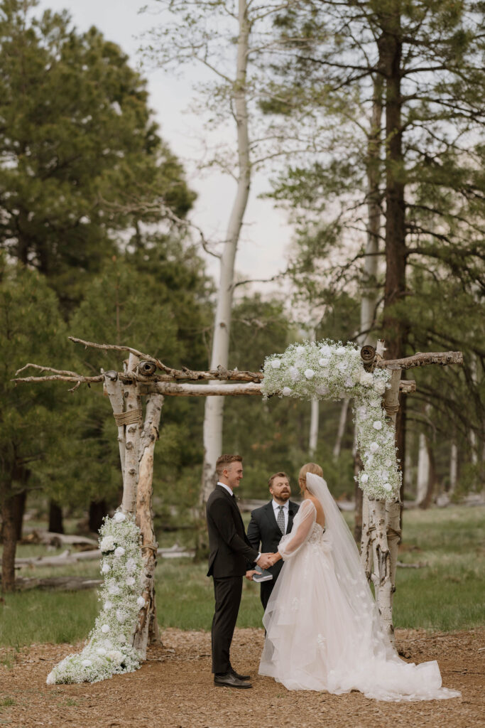 ceremony photo of groom holding hands at arbor with bride during outdoor wedding day at arizona nordic village